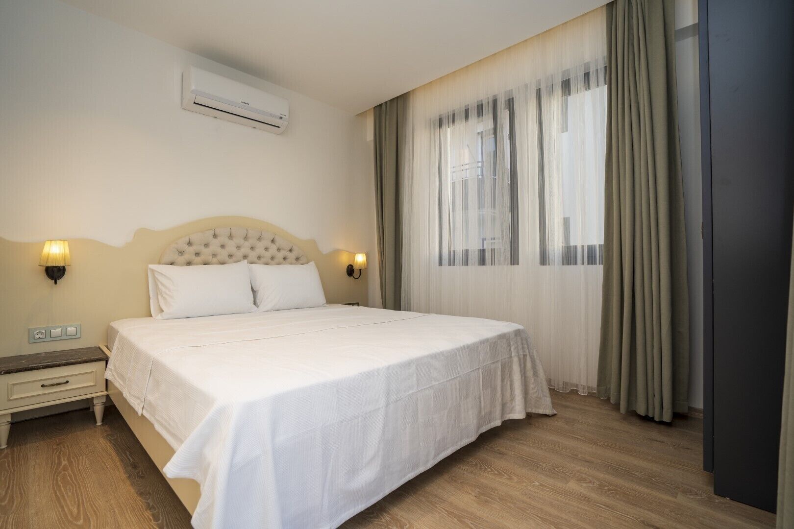 A hotel room with a large bed, two bedside lamps, an air conditioning unit, and a window with curtains.