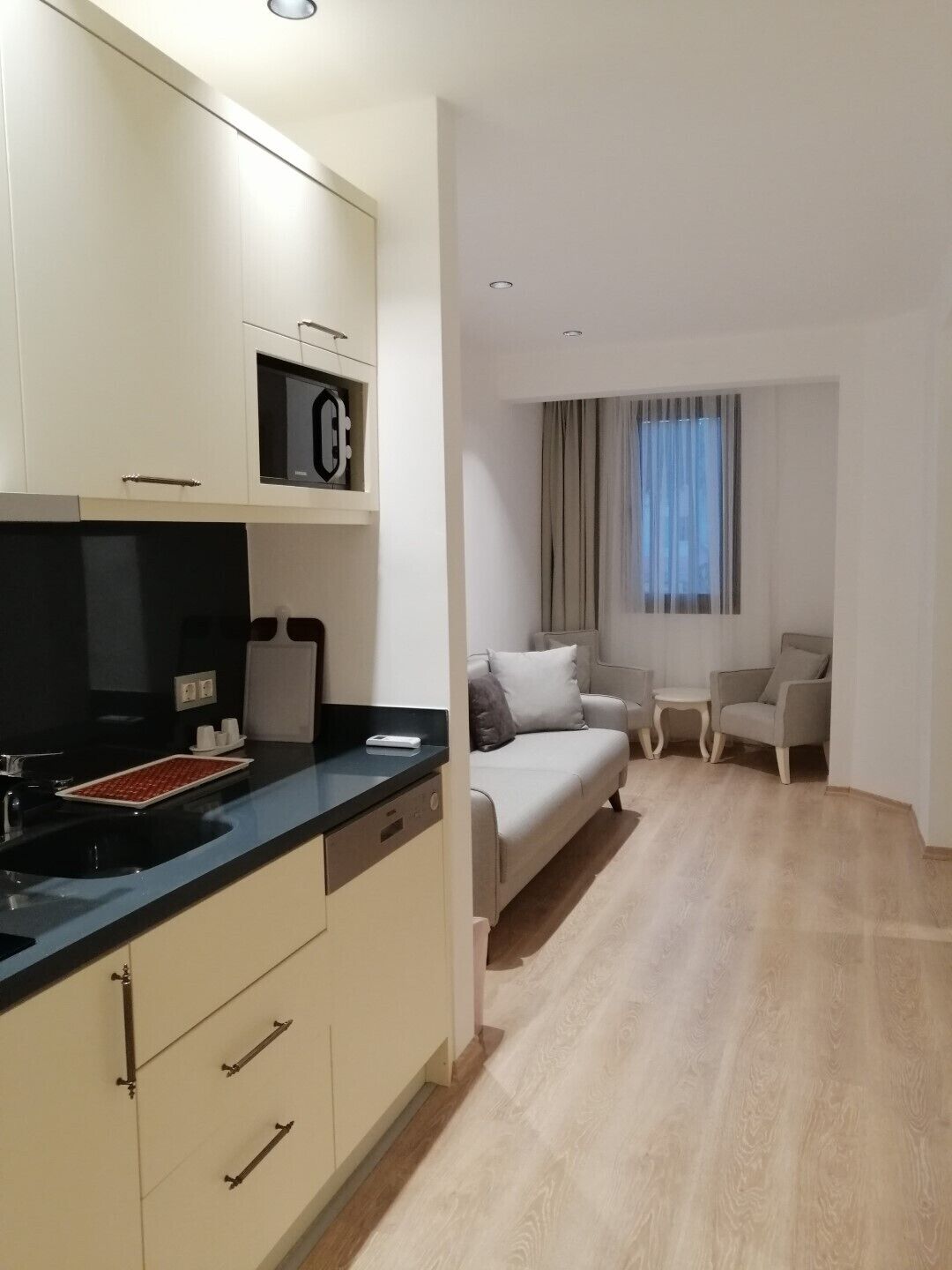 A modern kitchen with a black countertop, white cabinets, and a built-in microwave. There is a small dining area with a table and two chairs. The room has a large window with curtains and a light-colored wooden floor.