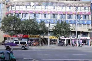 Un edificio de varios pisos con varios comercios en la planta baja, una zona peatonal con árboles y un vehículo estacionado.
