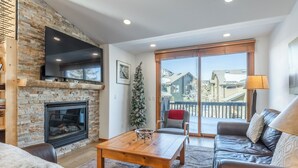 A cozy living room with a stone fireplace, a flat-screen TV, leather sofas, and a large window offering a view of a residential area.