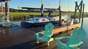 Un muelle de madera con dos sillas Adirondack verdes, un velero amarrado en el muelle y un edificio con un letrero con la letra 'M' al fondo.