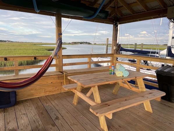 A wooden deck with picnic tables and a hammock overlooking a water body with boats.