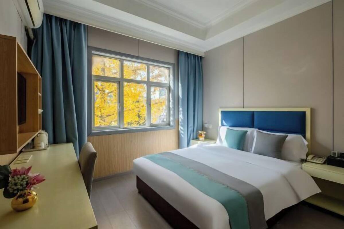 A hotel room with a large bed, a desk with a vase of flowers, a window with curtains, and a view of autumn trees.