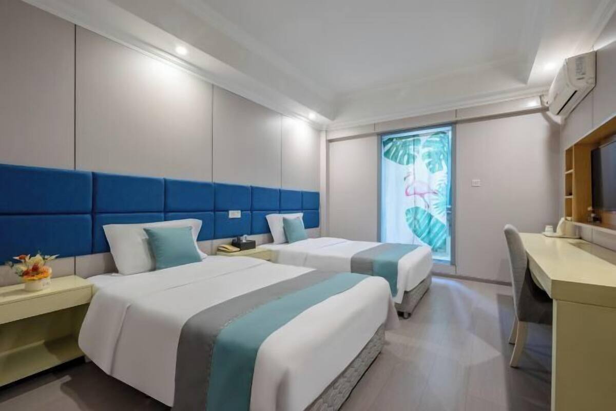 A hotel room with two beds, a desk, and a window with a view of greenery.