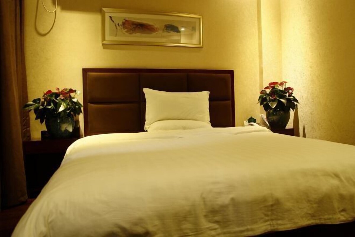 A hotel room with a large bed, a headboard, a framed picture on the wall, and two potted plants on the nightstand.