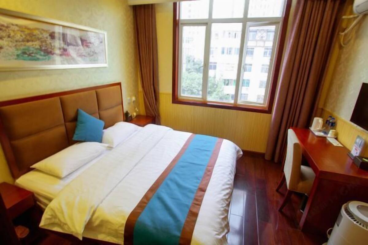 A hotel room with a bed, a desk, a chair, a window with curtains, and a painting on the wall.