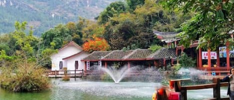 A traditional Chinese water pavilion with a wooden walkway, surrounded by lush greenery and a mountainous backdrop.
