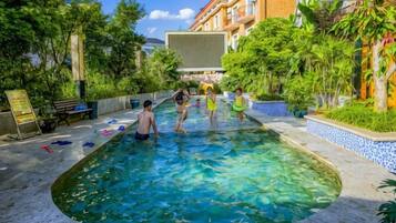 Una piscina con gente a su alrededor, rodeada de vegetación y un edificio.