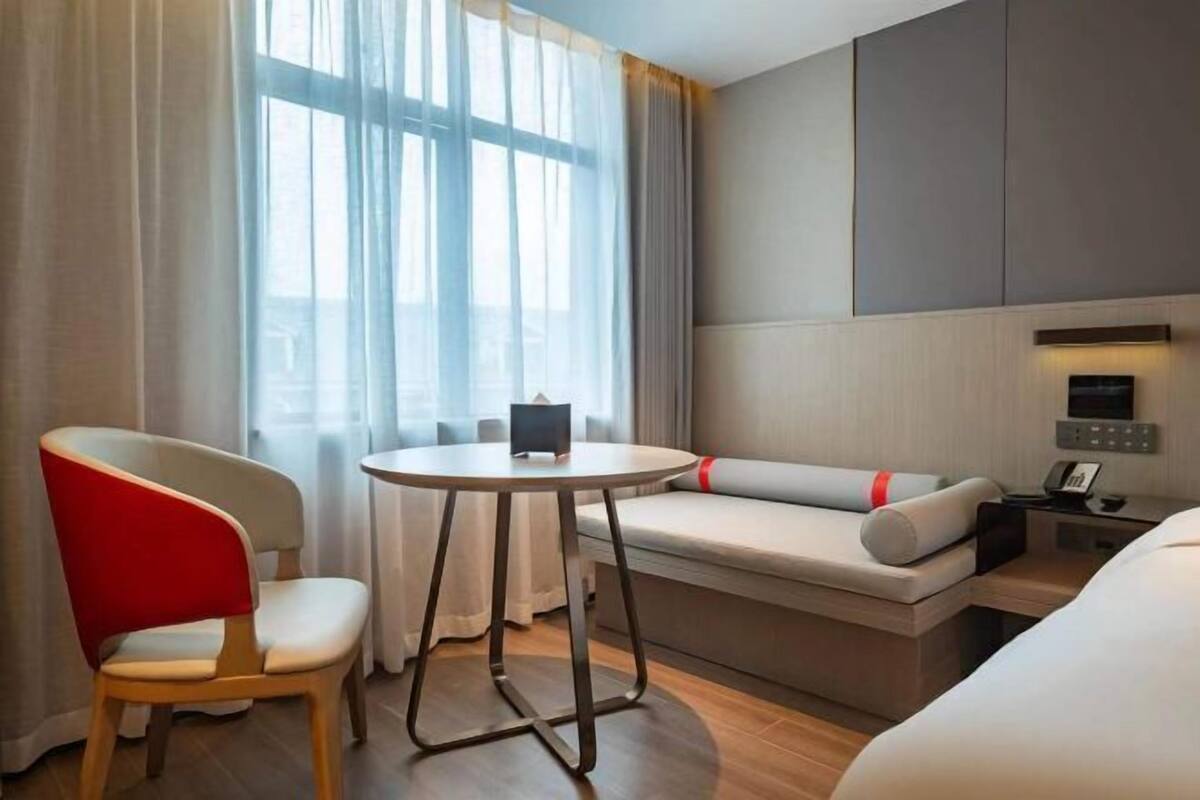 Ein Hotelzimmer mit einem Bett, einem roten Sessel, einem kleinen Tisch, einer Couch und einem Fenster mit Vorhängen.