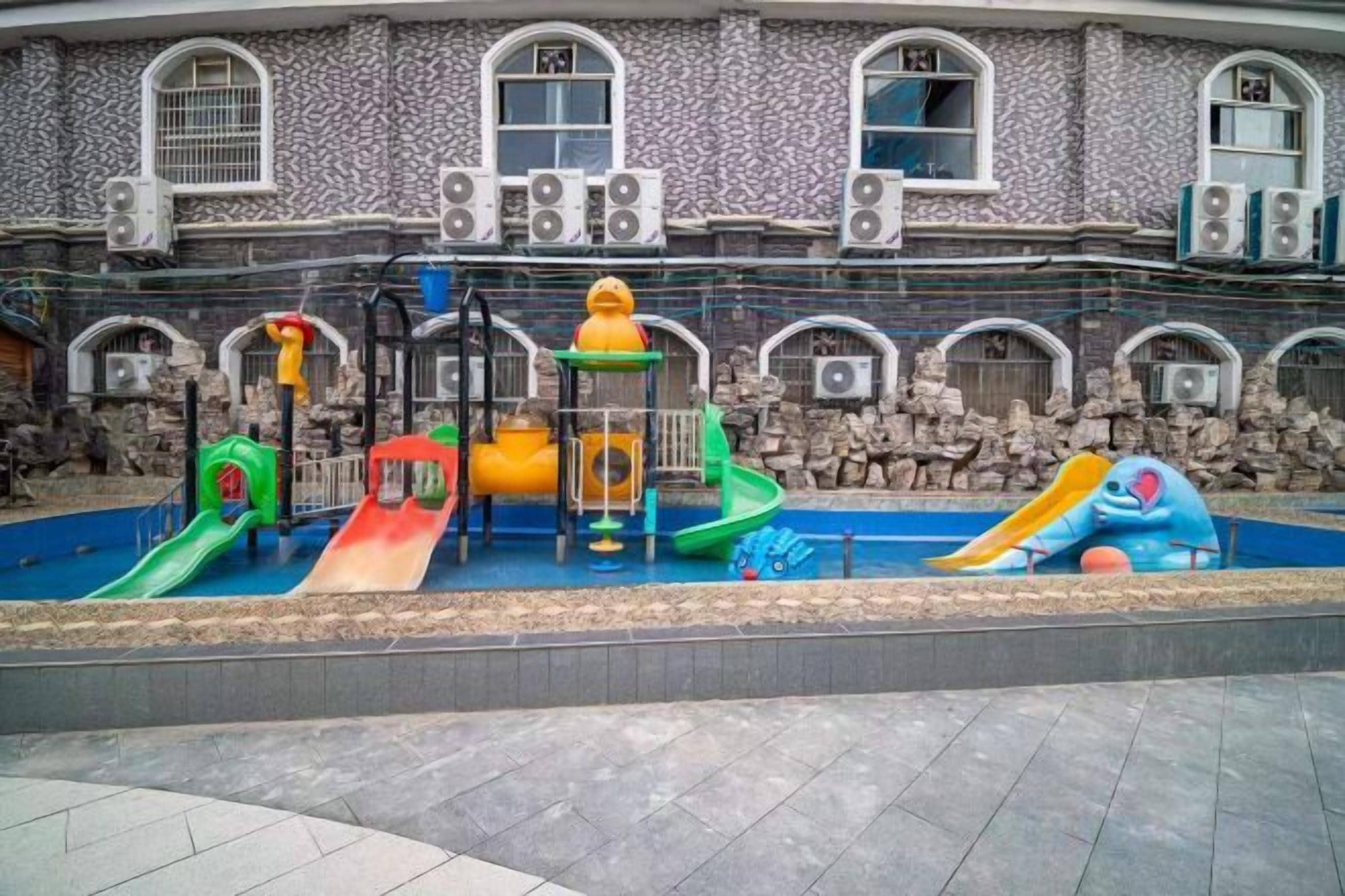 Un parque infantil con toboganes y estructuras de escalada frente a un edificio de paredes de piedra y ventanas de arco.