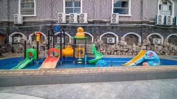 Un parque infantil con toboganes y estructuras de escalada frente a un edificio de paredes de piedra y ventanas de arco.