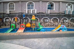Ein Spielplatz mit mehreren Rutschen und Klettergerüsten vor einem Gebäude mit Rundbogenfenstern und Steinmauern.