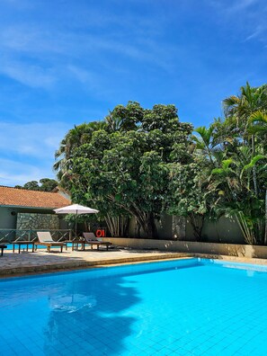 A swimming pool with lounge chairs and umbrellas, surrounded by lush greenery and a stone wall. - Pantanal Hotel (Miranda)