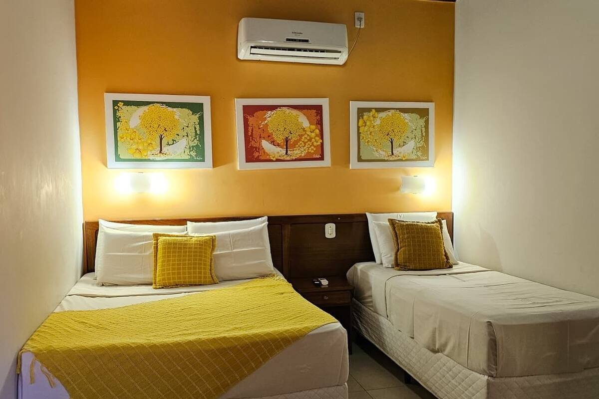 Habitación de hotel con dos camas, aire acondicionado de pared y tres cuadros con imágenes de árboles.