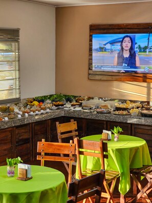 A breakfast buffet with various food items, a television displaying a news broadcast, and a hotel sign on a table. - Pantanal Hotel (Miranda)