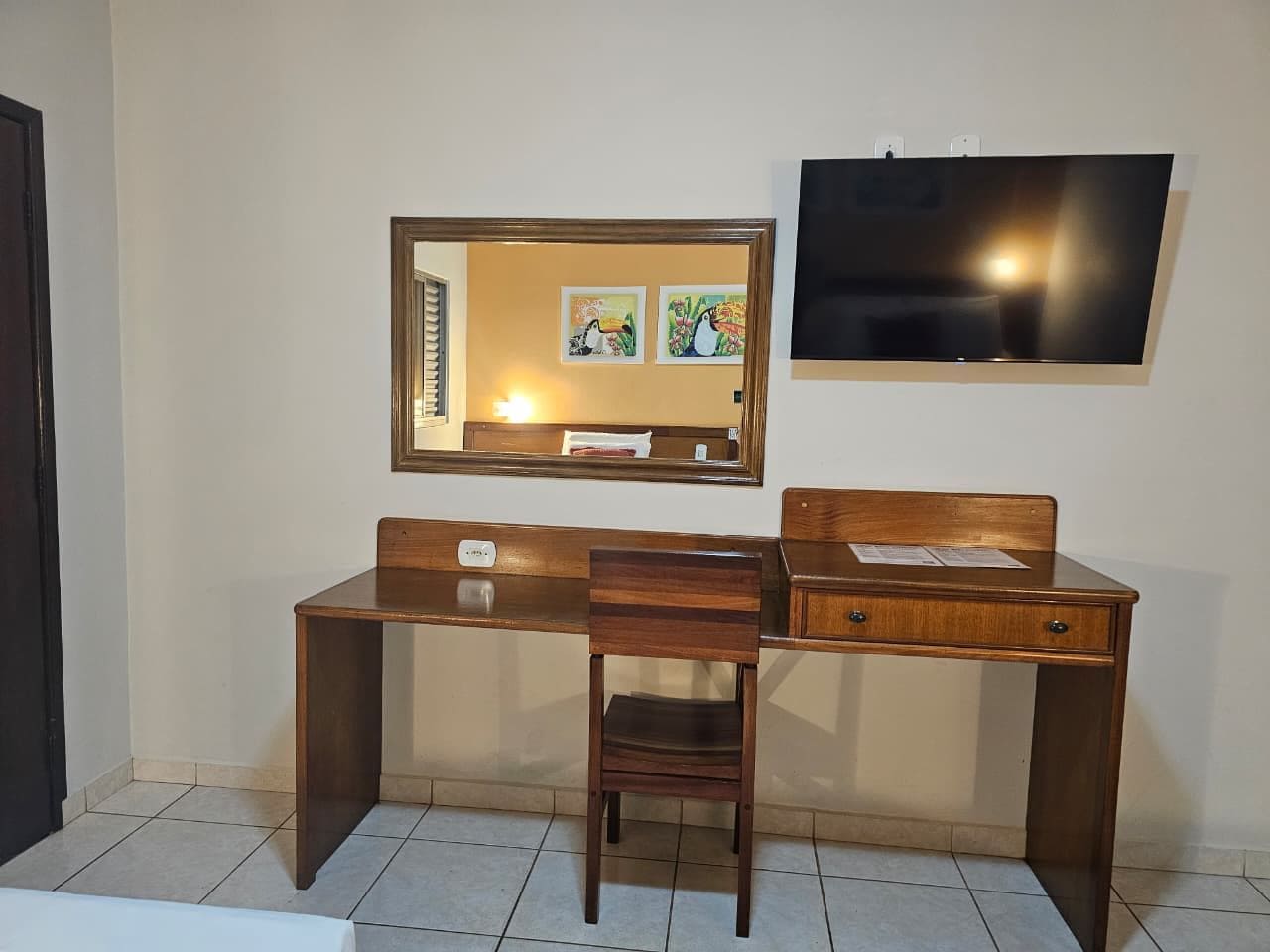 Quarto com escrivaninha de madeira, cadeira, espelho e televisão de tela plana fixada na parede.