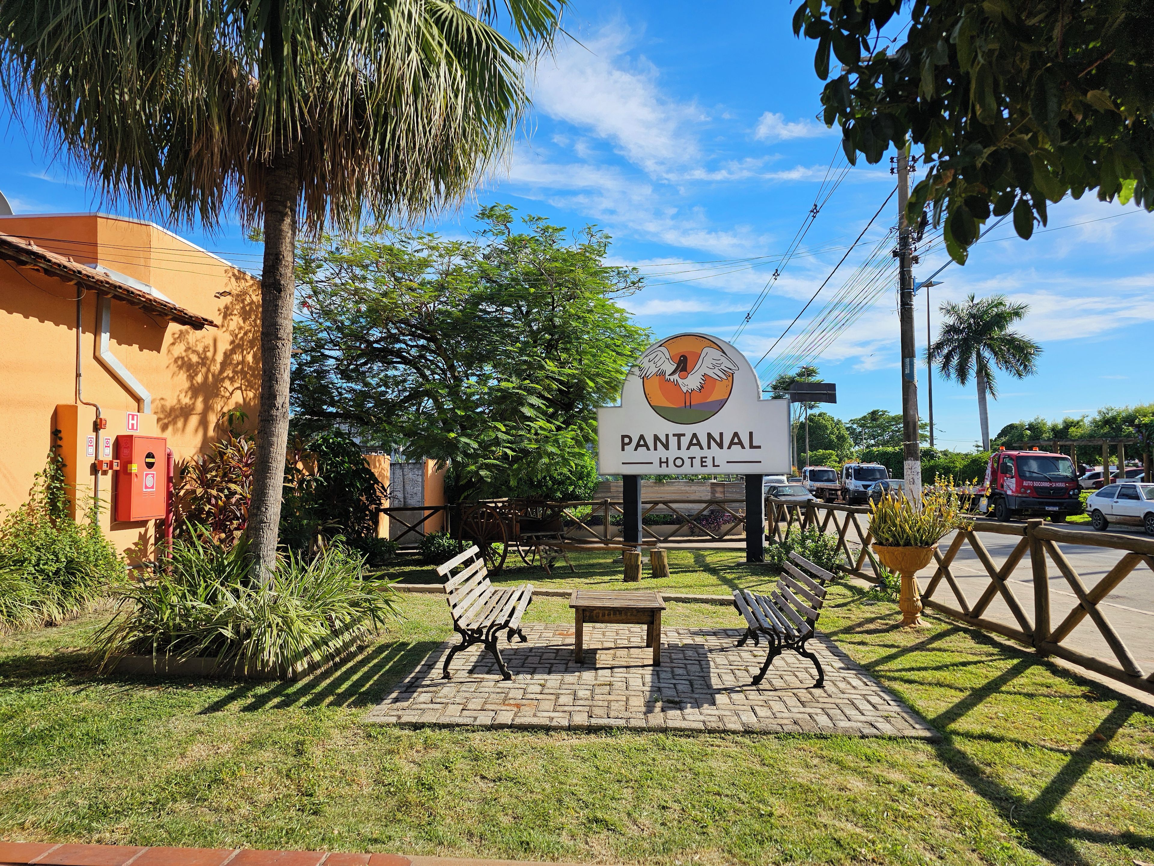 Placa do Hotel Pantanal com logotipo, cercada por vegetação e área pavimentada com bancos.