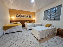 Quarto de hotel com quatro camas, cabeceira de madeira, ar-condicionado fixado na parede e janela com persianas.