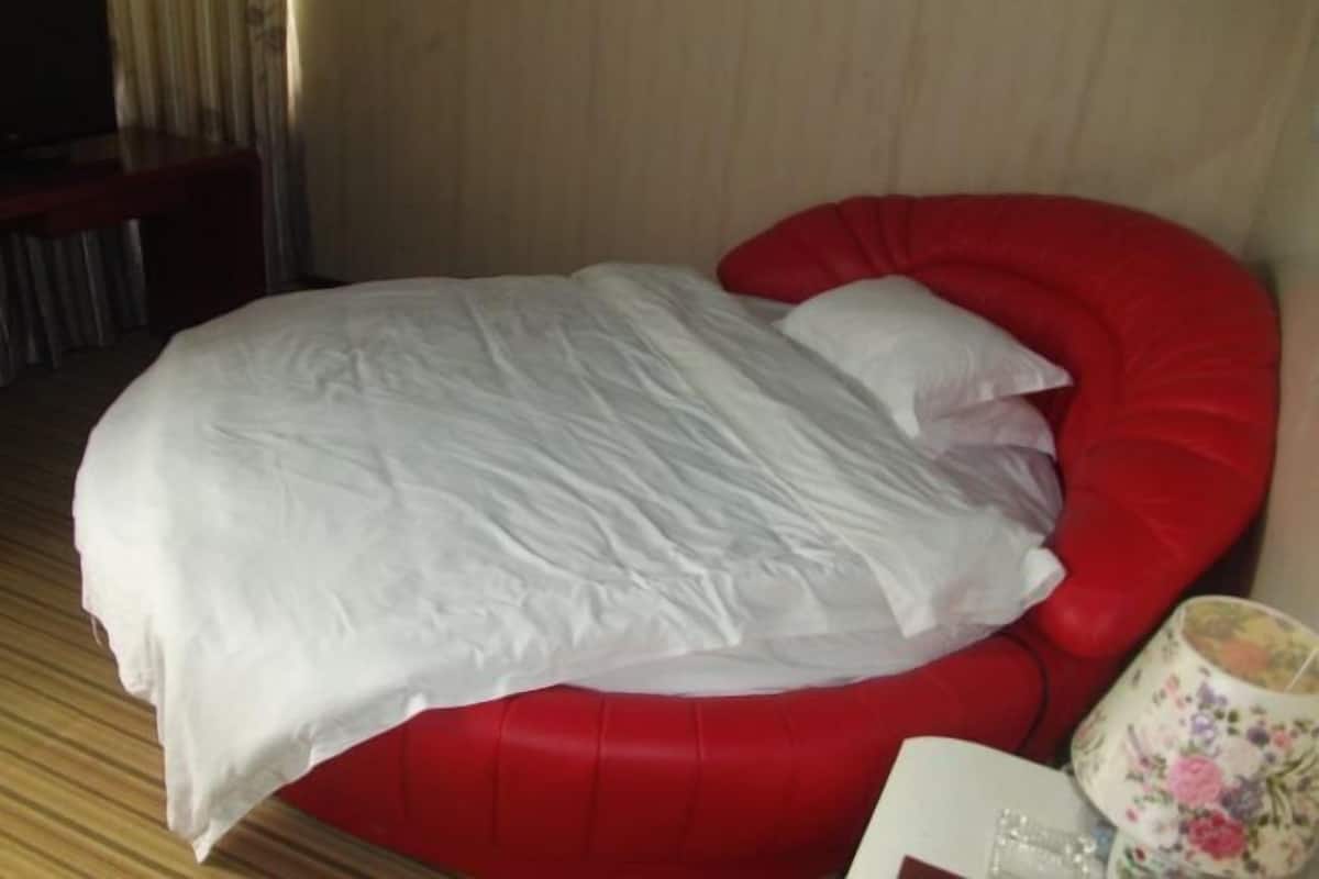 Een bed met een witte dekbedovertrek en kussens, een rode beklede stoel, een lamp met bloemenprint en een houten vloer.