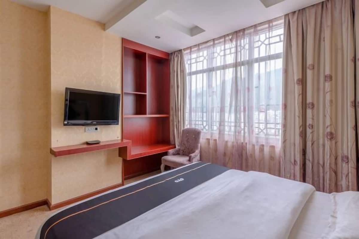 Hotelkamer met een bed, televisie, inbouwkasten, een stoel en grote ramen met gordijnen.