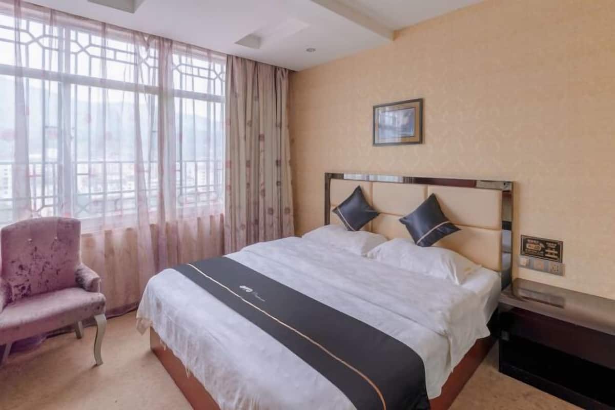 Een hotelkamer met een bed, een stoel, een raam met gordijnen en een lijst met een afbeelding aan de muur.