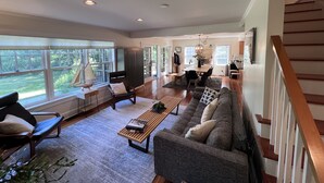 A spacious living room with a large sofa, a coffee table, and a dining area with a table and chairs. There is a staircase on the right side and large windows on the left.