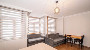 Quarto com cama, sofá, escrivaninha com cadeiras e janelas amplas com persianas.