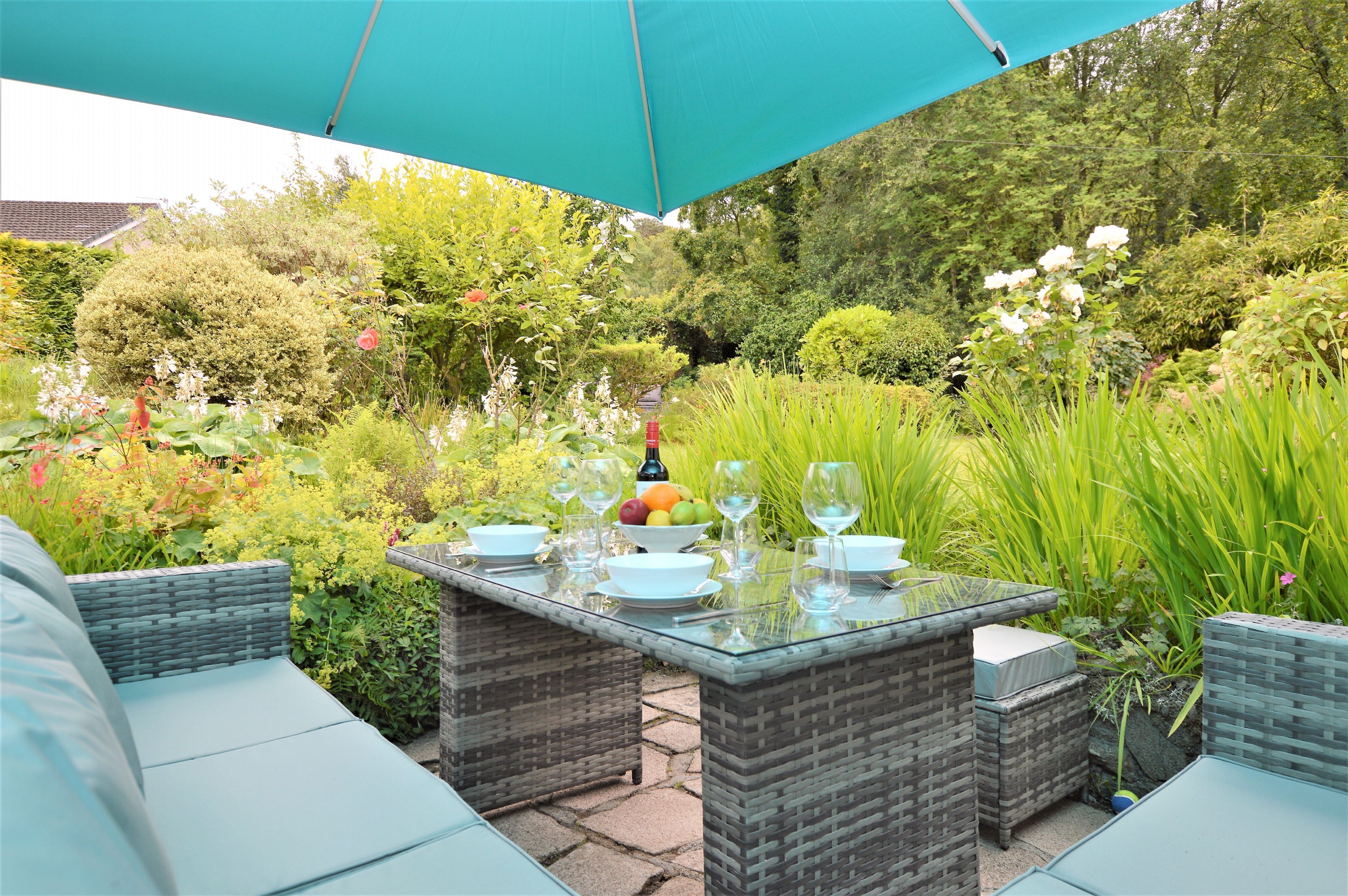 An outdoor dining area with wicker furniture and a glass table, surrounded by lush greenery and a large umbrella.
