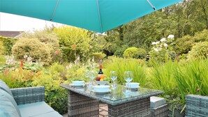 An outdoor dining area with wicker furniture and a glass table, surrounded by lush greenery and a large umbrella. - Brookfield, picturesue quiet location, sleeps 8, pet friendly (Dunoon)