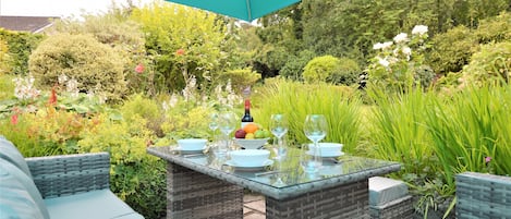 An outdoor dining area with wicker furniture and a glass table, surrounded by lush greenery and a large umbrella.