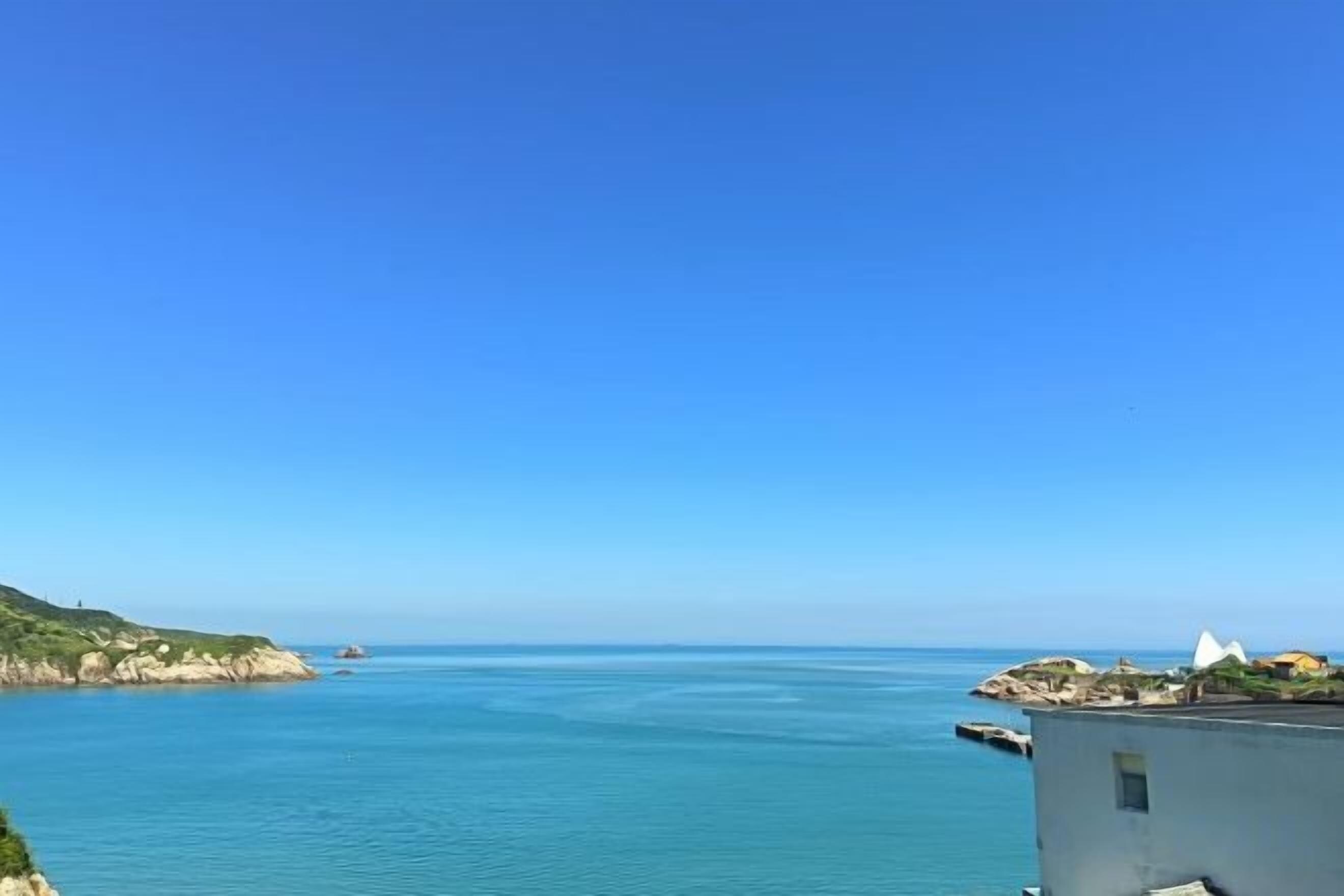 A coastal view with a clear sky, calm sea, and a building with a flat roof and a small balcony.