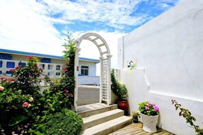 A white building with a wooden staircase leading to an arched entrance, surrounded by potted plants and flowers.
