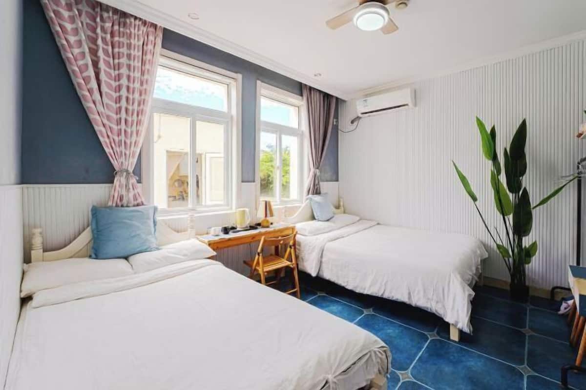 Een kamer met twee bedden, een bureau, een stoel, een kamerplant en een raam met gordijnen.