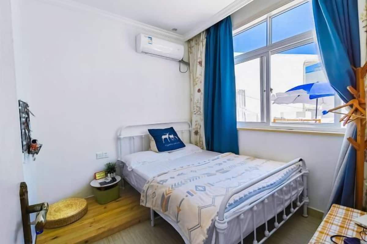 Een kleine, verzorgde slaapkamer met een enkel bed, een raam met gordijnen, een nachtkastje en een wandairconditioner.