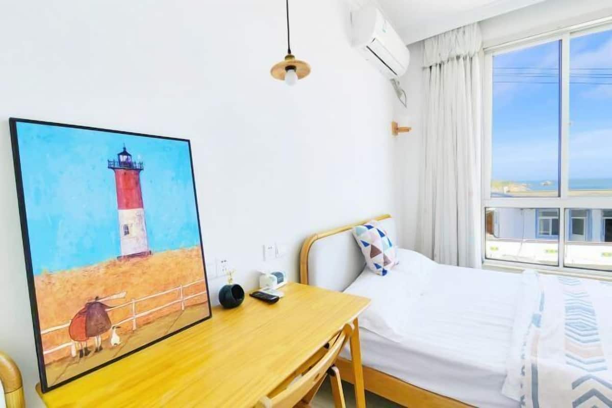 Hotelkamer met een bed, een bureau met stoel, een schilderij van een vuurtoren, een airconditioner en een raam met uitzicht.