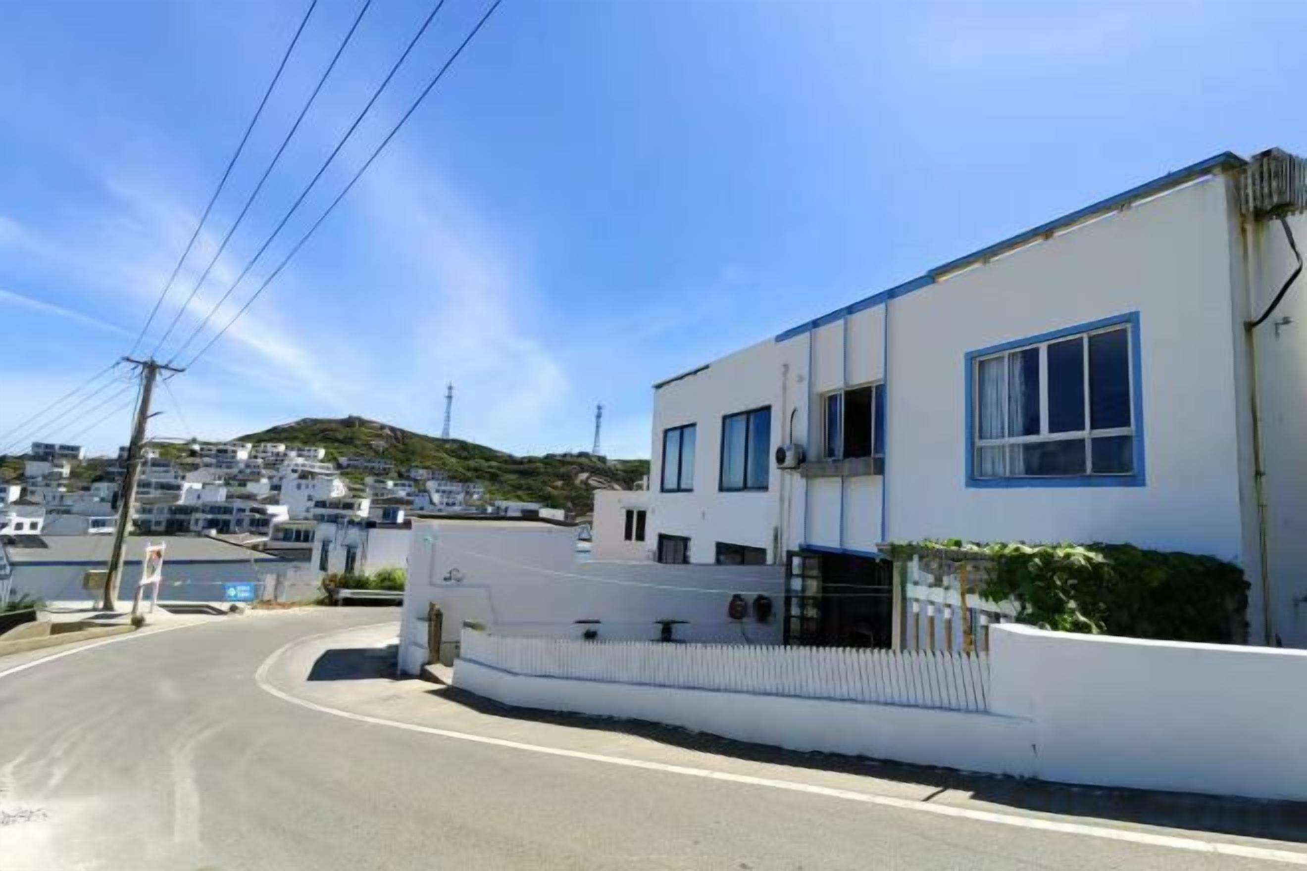 A residential street with modern houses, a clear sky, and a hilly background.