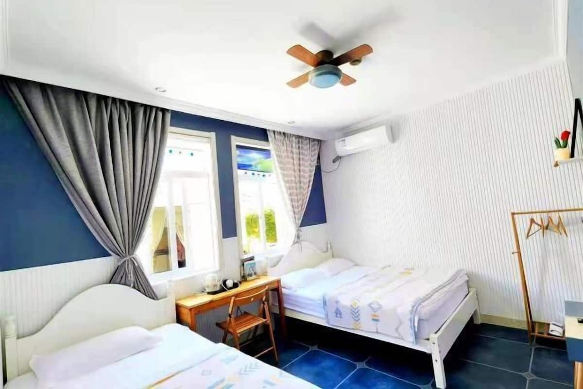 Een kamer met twee bedden, een plafondventilator, een raam met gordijnen, een bureau en een stoel.
