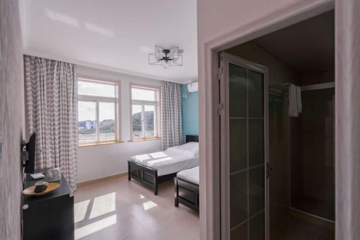 Een slaapkamer met twee bedden, een bureau, een stoel, een raam met gordijnen, een plafondventilator en een glazen deur naar een badkamer.