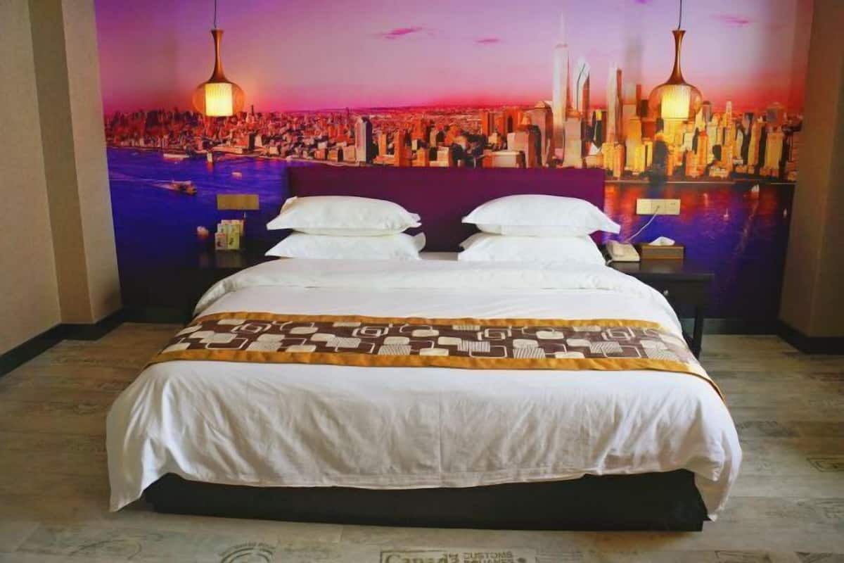 A hotel room with a bed, a nightstand, and a wall mural of a cityscape at sunset.