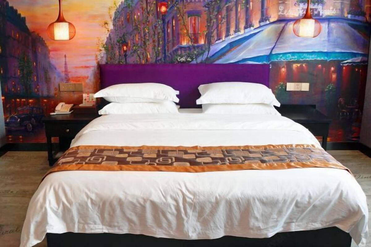 A hotel room with a large bed, purple headboard, and a cityscape mural on the wall.