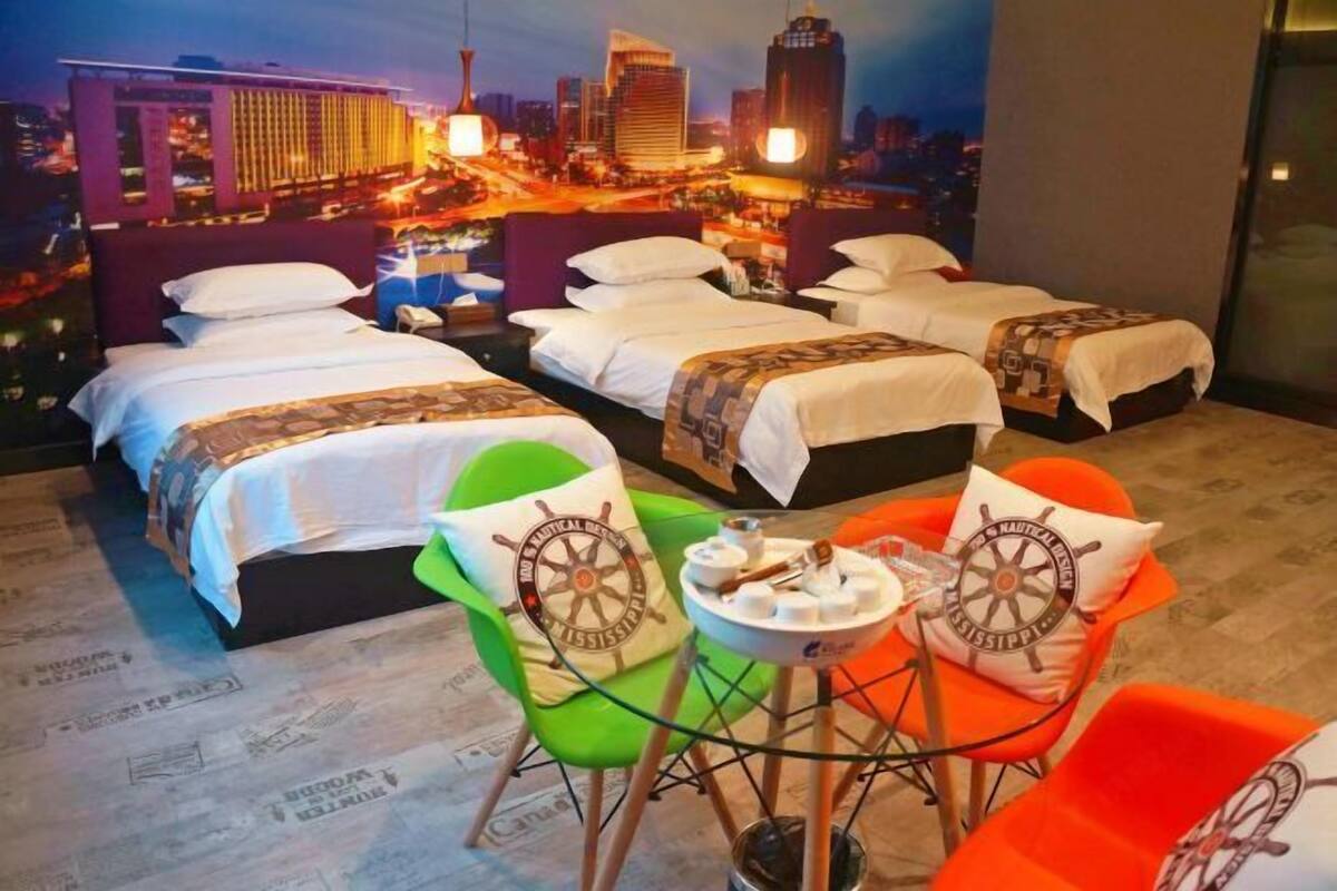 A hotel room with three beds, a cityscape mural, a small table with a chair, and nautical-themed pillows.