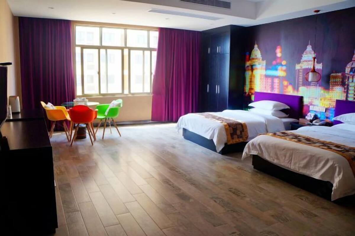 A hotel room with two beds, a desk, chairs, and a large window with curtains.