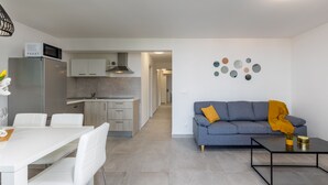 A modern kitchen with white cabinets, a grey refrigerator, and a microwave. There is a dining table with white chairs and a grey sofa with a yellow throw blanket. A coffee table is in front of the sofa. - Luxurious apartment D (Crikvenica)