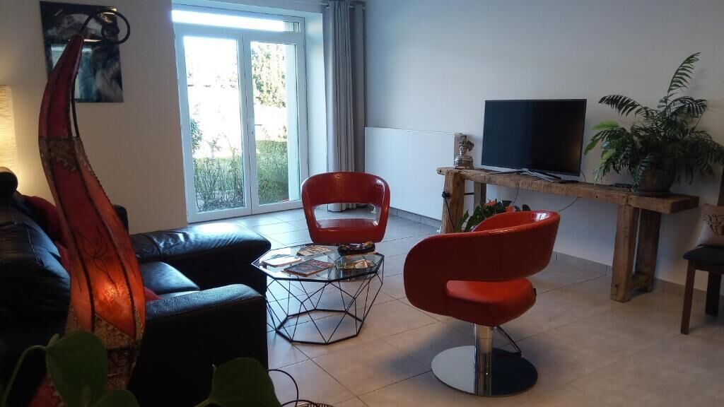 Een moderne woonkamer met een bank, twee rode stoelen, een glazen salontafel, een tv op een houten dressoir, en een groot raam met uitzicht op groen.