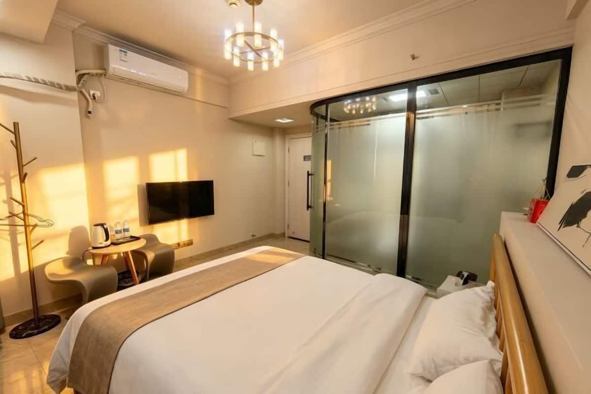 A hotel room with a bed, a desk with a chair, a TV, and a glass door leading to a bathroom.