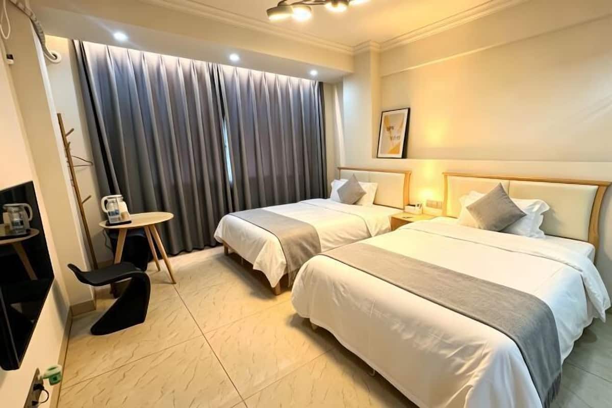 A hotel room with two beds, a small table with a chair, a coffee machine, and a large window with curtains.