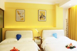 A hotel room with two beds, each with a heart-shaped pillow, white bedding, and a small bedside table with a lamp.