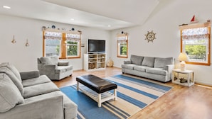 A spacious living room with two grey sofas, a black bench, a flat-screen TV, and a ceiling fan. - Best Kept Secret: Soundside, Hot Tub, Dogs OK (Kill Devil Hills)