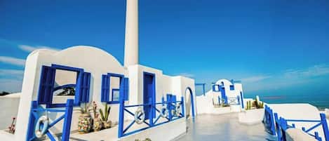 A coastal building with white walls and blue accents, featuring a staircase leading to a terrace with a view of the sea.