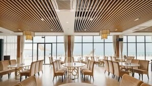 Un comedor amplio con mesas y sillas redondas, ventanas grandes con vistas al mar y un techo con listones de madera.